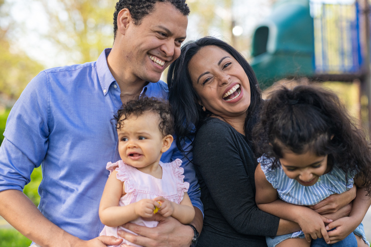 A happy Latin American family is spending time together. A mother and father are embracing their daughters while outdoors. The family is posing and laughing towards the camera. It's a warm, sunny day. A playground is visible in the background.